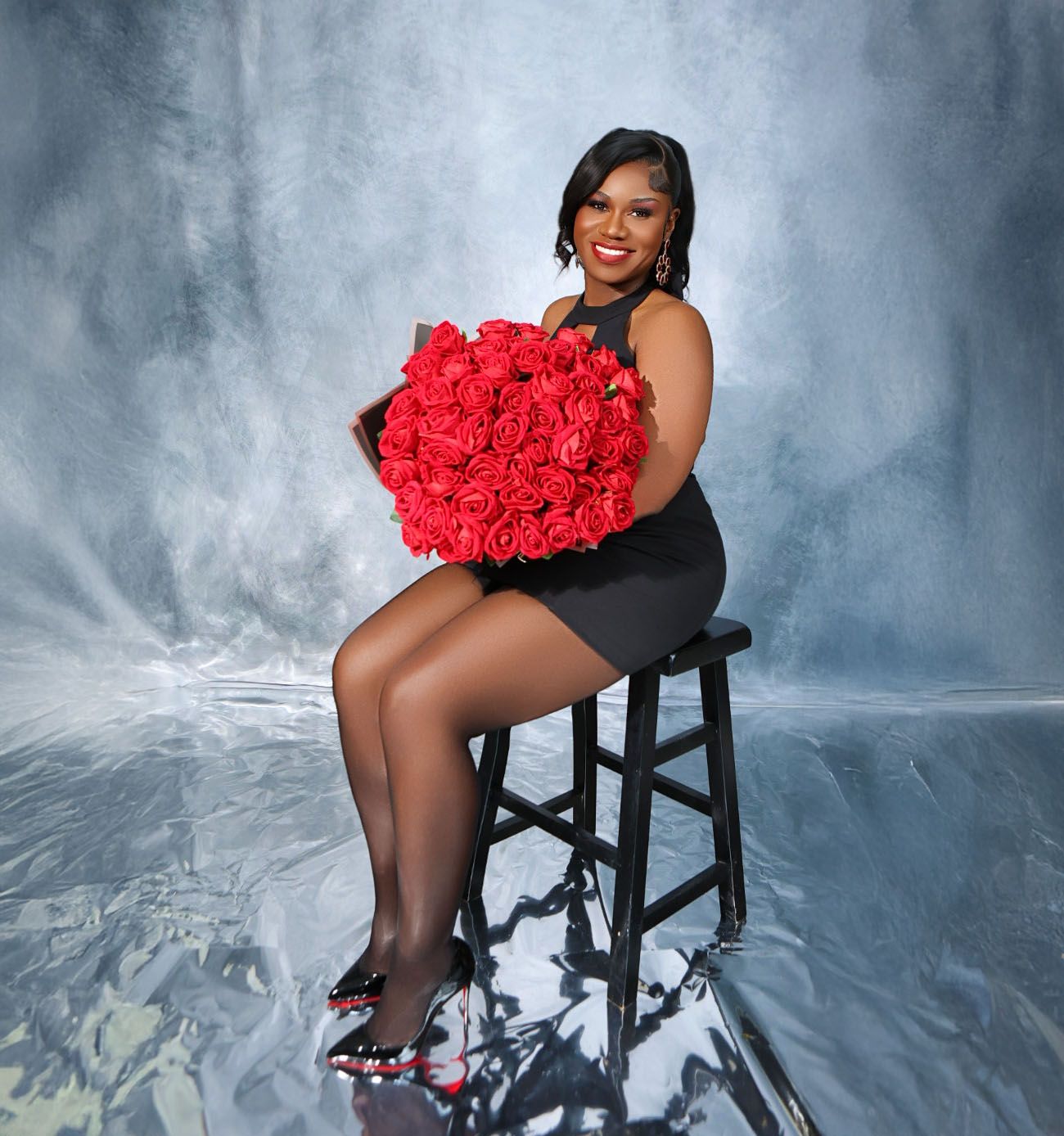 Smiling woman in black dress sitting on stool holding large bouquet of red roses in studio.