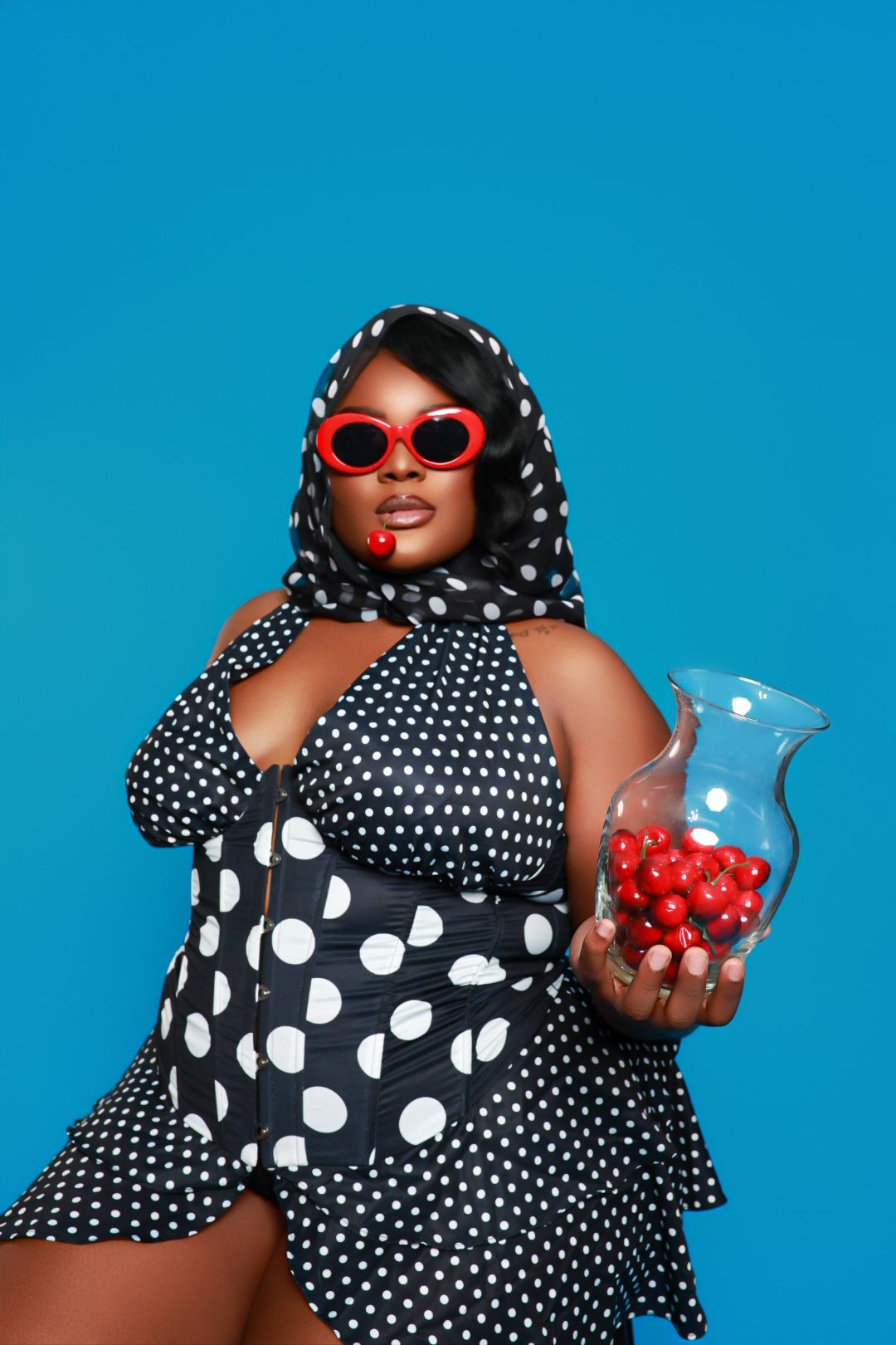 Woman in polka dot outfit holding cherries, posing against a blue background.
