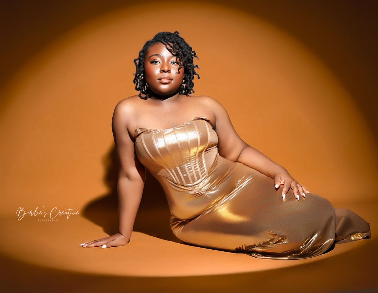 Woman in gold metallic strapless gown posing elegantly on floor against warm amber background.