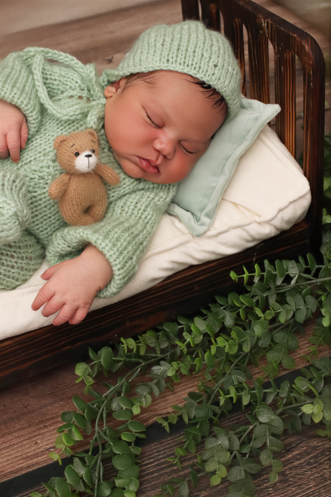 Newborn baby sleeping in green knit outfit with teddy bear in a tiny wooden bed.