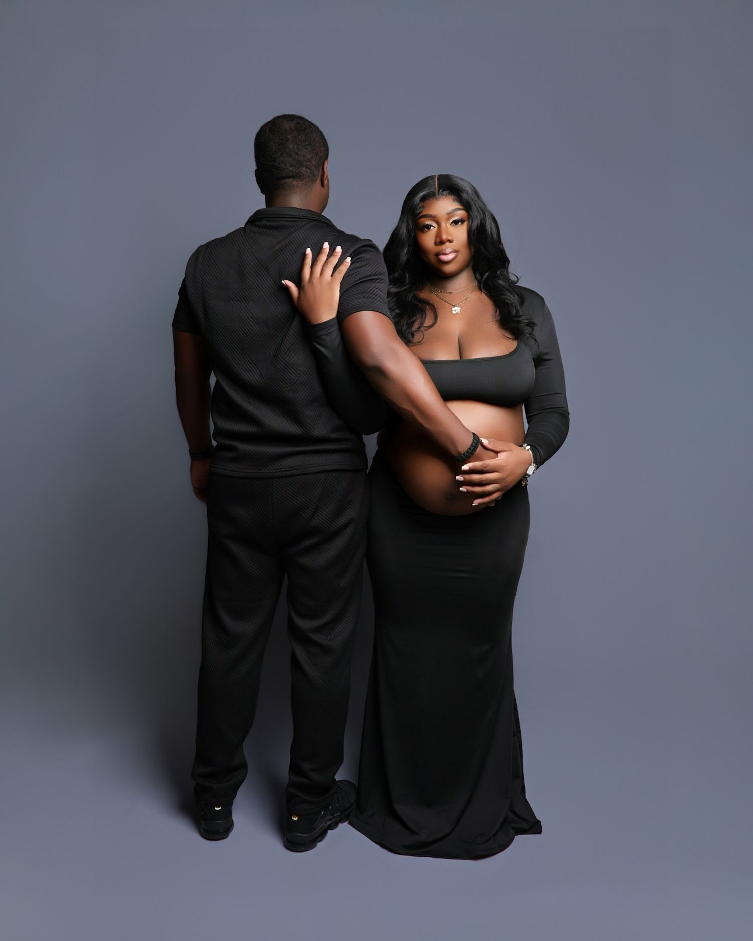 Maternity couple photo in black outfits, woman in elegant gown cradling baby bump on gray backdrop.