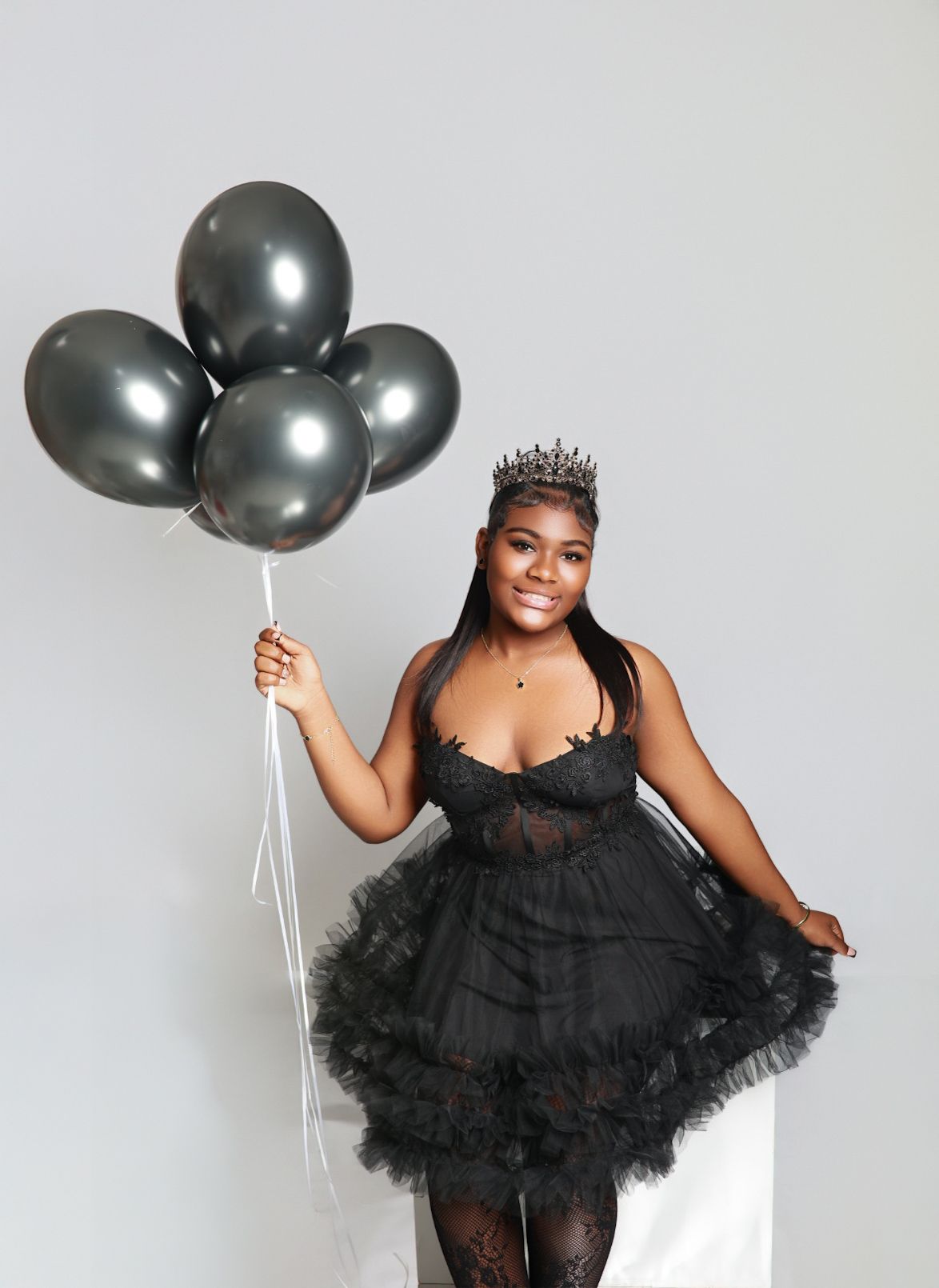 Woman in black tulle dress and crown holds silver balloons against white background.