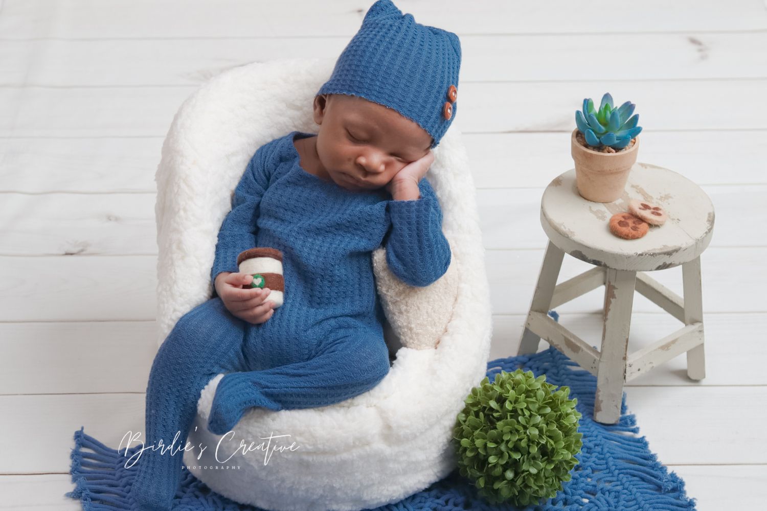 Newborn baby in blue knit outfit sits in white chair holding small jar in studio portrait.