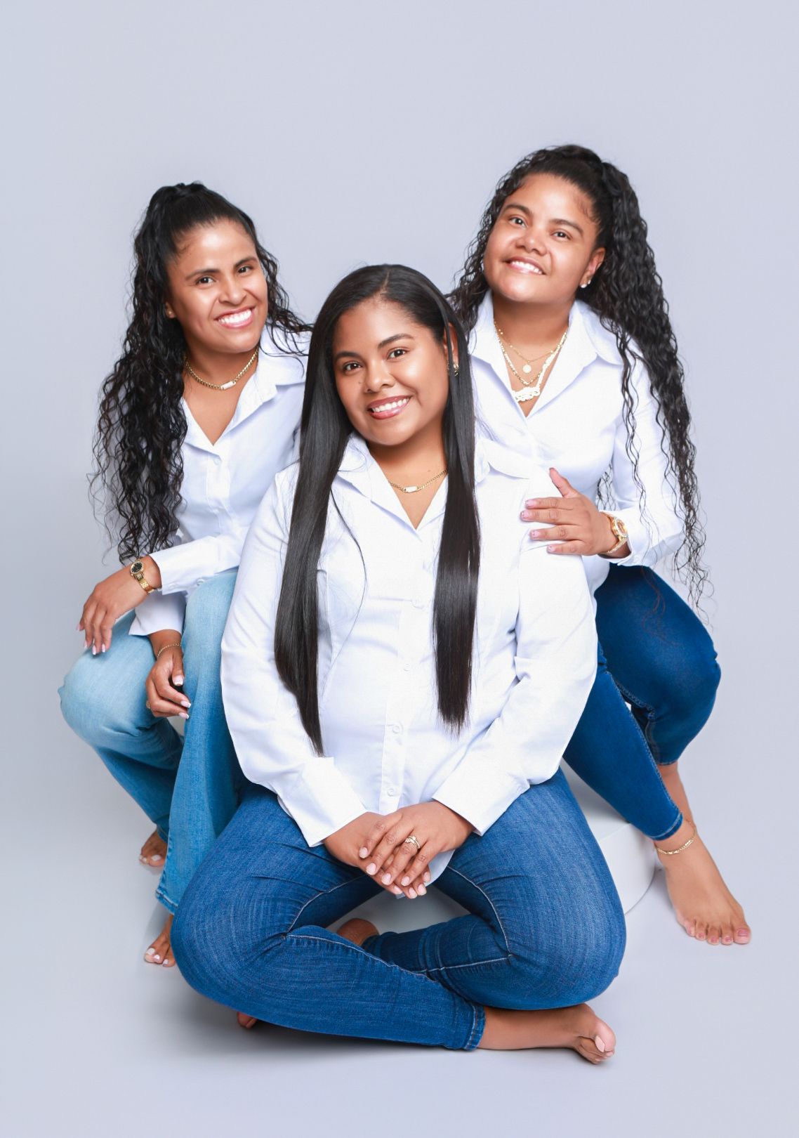 Three women in white shirts and jeans pose together in a close-knit studio portrait on the floor.