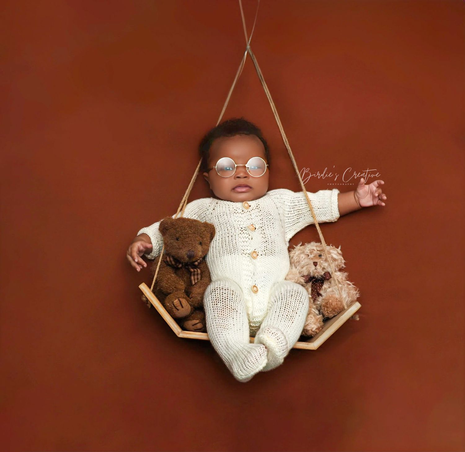 Newborn baby in white knit outfit with tiny sunglasses sitting in a hanging swing with teddy bears.