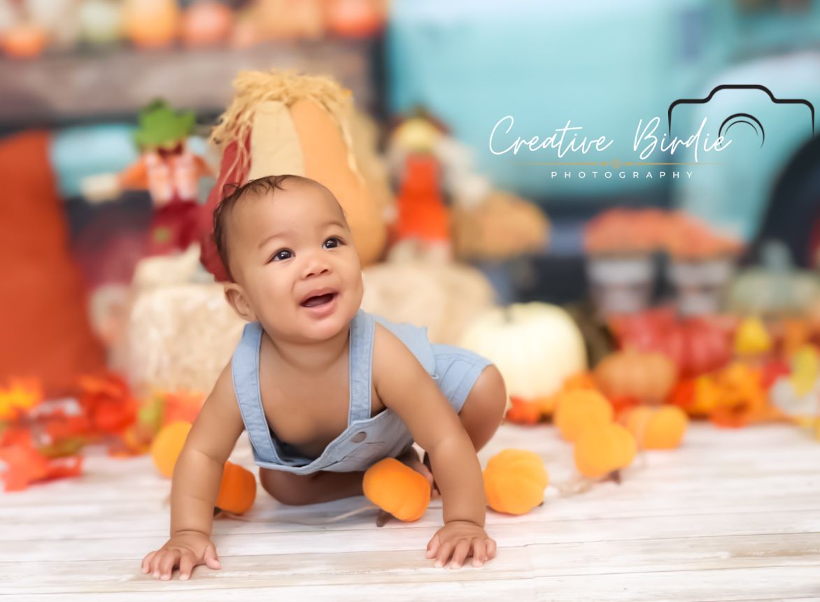 Happy smiling baby crawls on floor wearing fall harvest crown surrounded by colorful pumpkins.