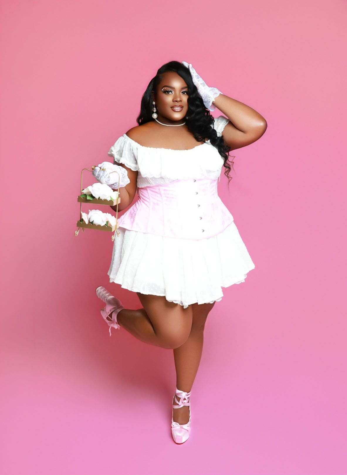 Plus-size woman in white corset mini dress holding bouquet poses against pink background.