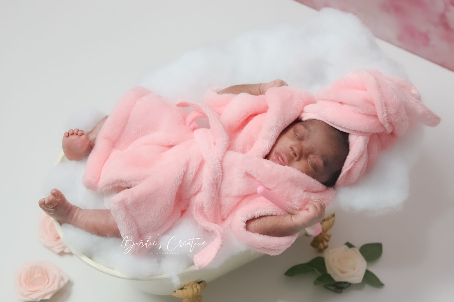 Newborn baby sleeping in pink fluffy robe in a small white bathtub with roses.