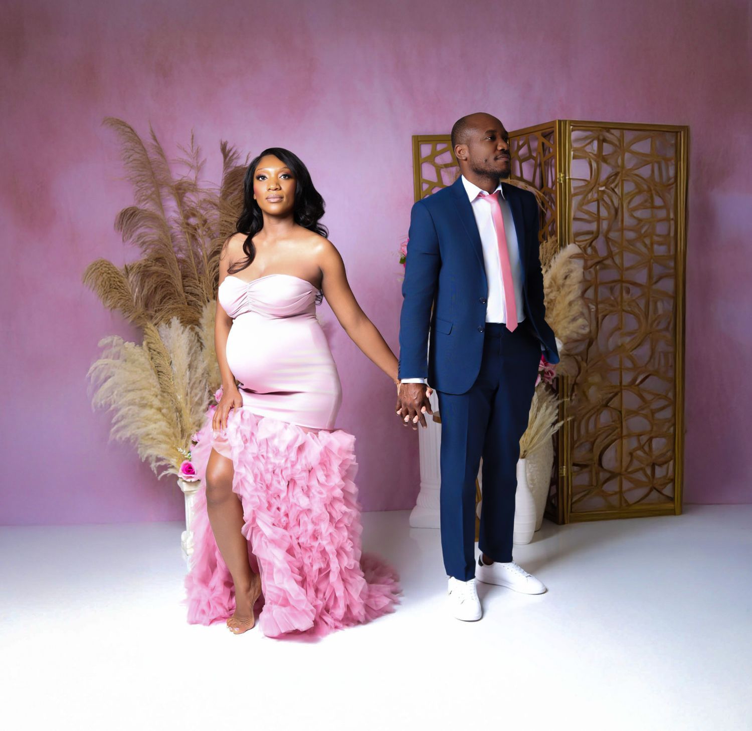 Maternity couple holding hands in pink studio, woman in ruffled pink gown and man in navy suit.