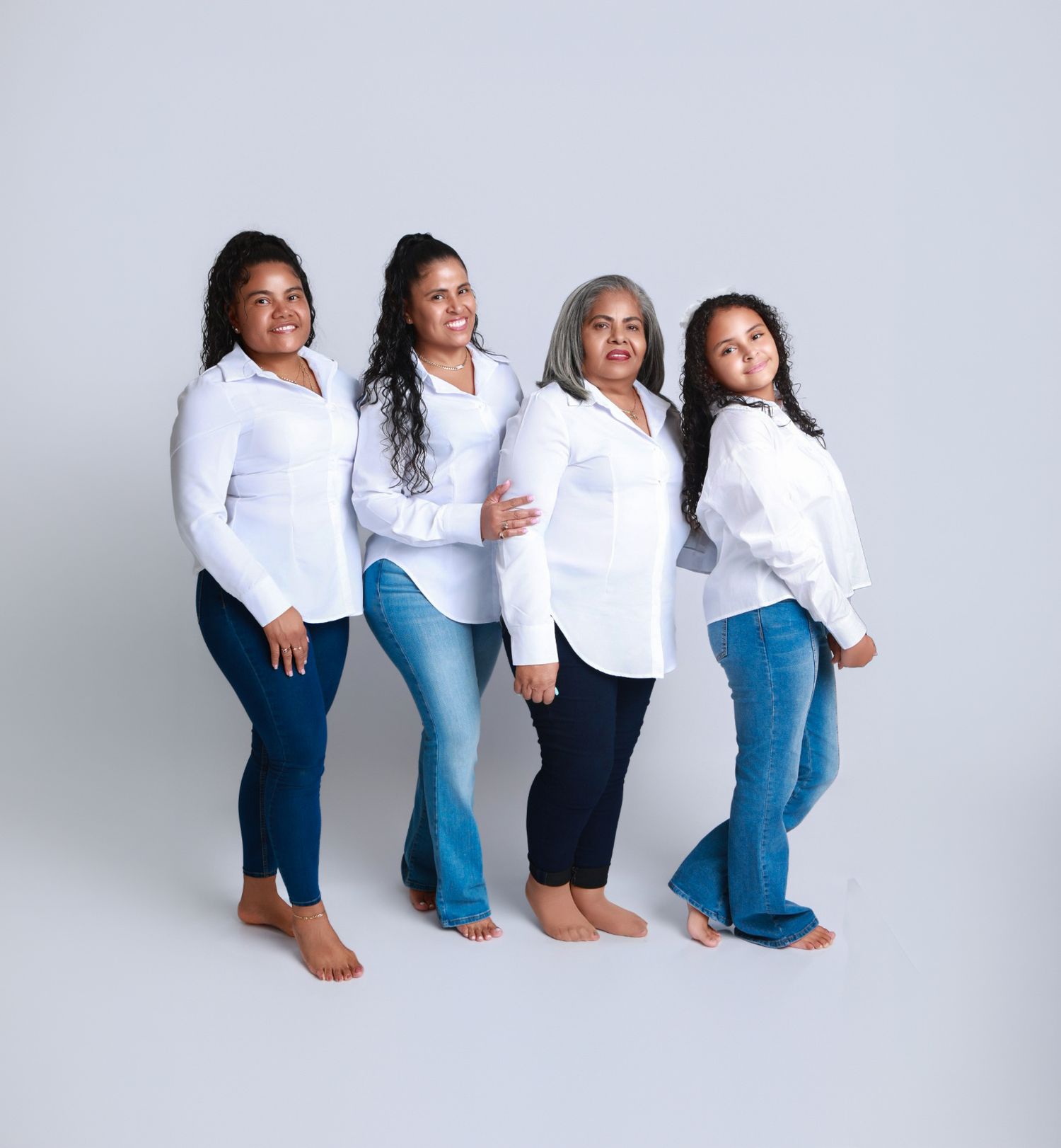 Four women wearing coordinated white button-up shirts and blue jeans pose standing in a studio portrait.