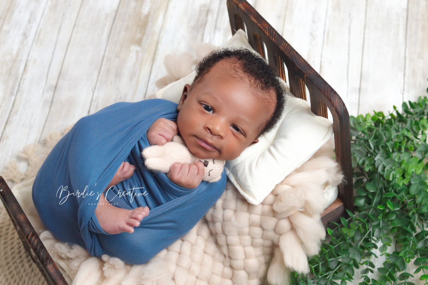 Newborn baby wrapped in blue swaddle holds white toy bear in rustic wooden bed photo prop.
