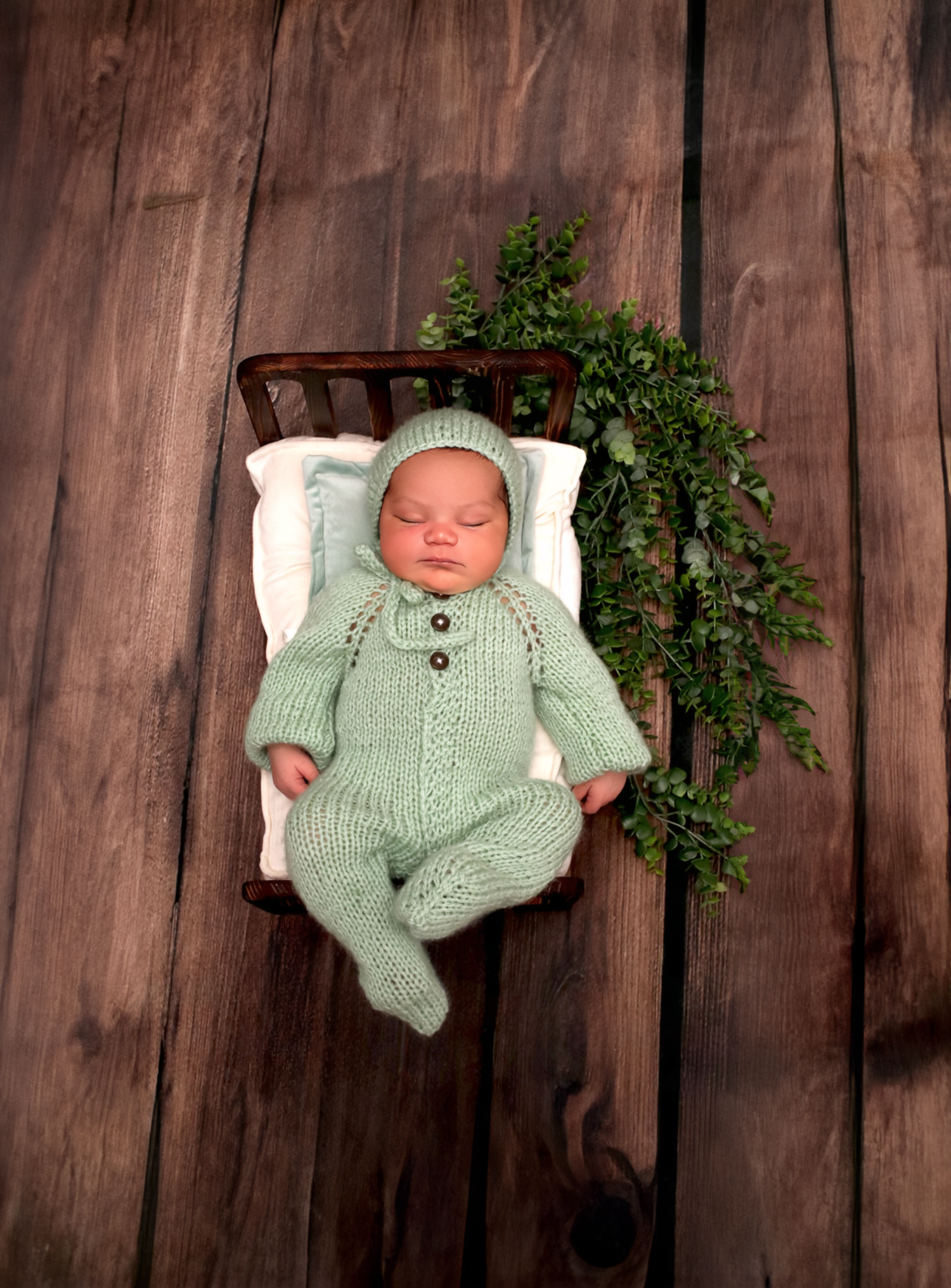 Sleeping newborn in mint green knit romper and hat in a small wooden bed.