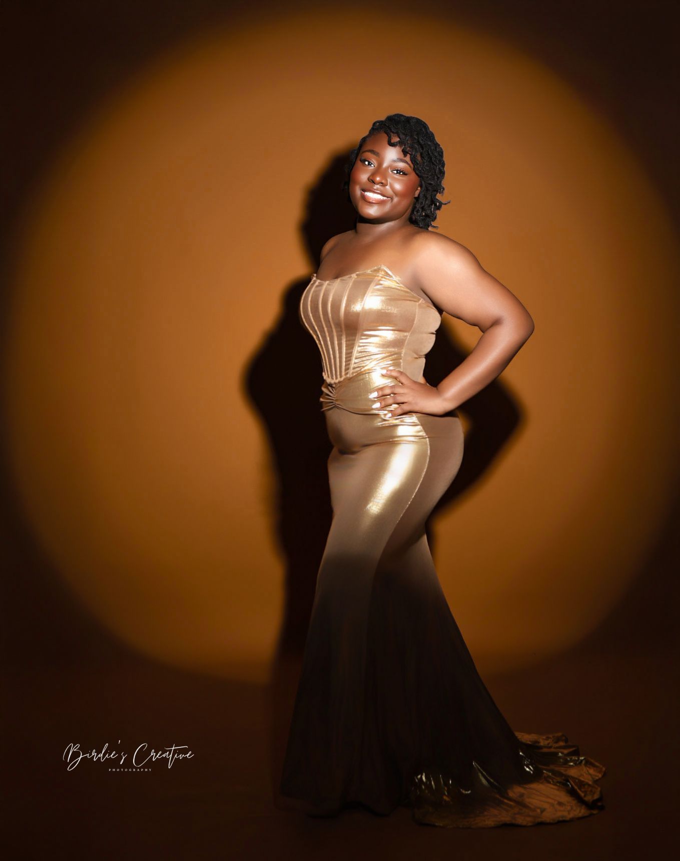 Woman in stunning gold ombre mermaid gown posing confidently against orange studio backdrop.
