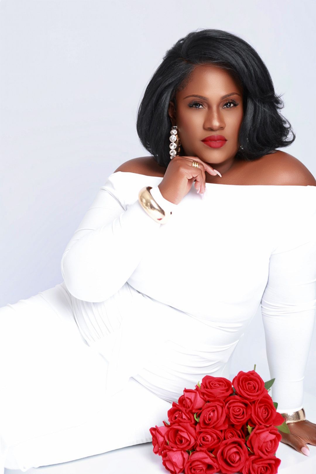 Woman in white off-shoulder dress reclining elegantly with red roses in professional studio portrait.