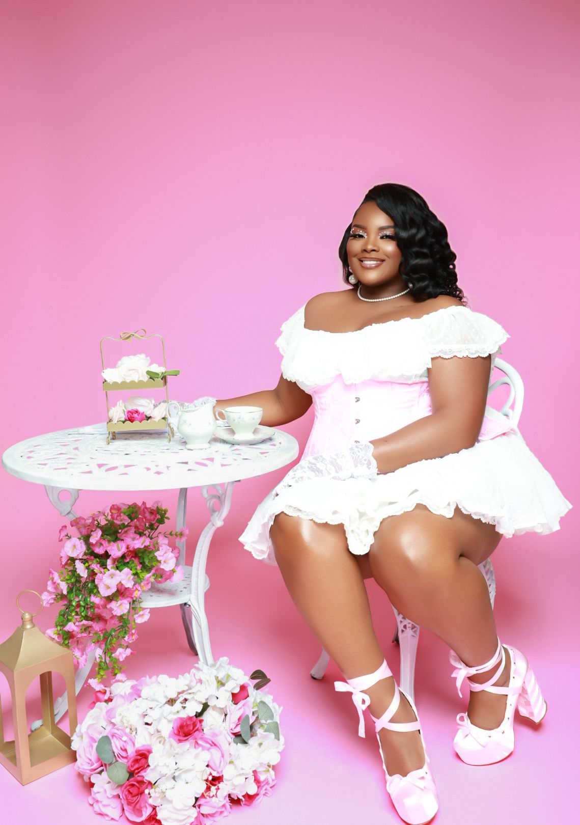 Woman in white ruffled outfit seated at a table on a pink background.