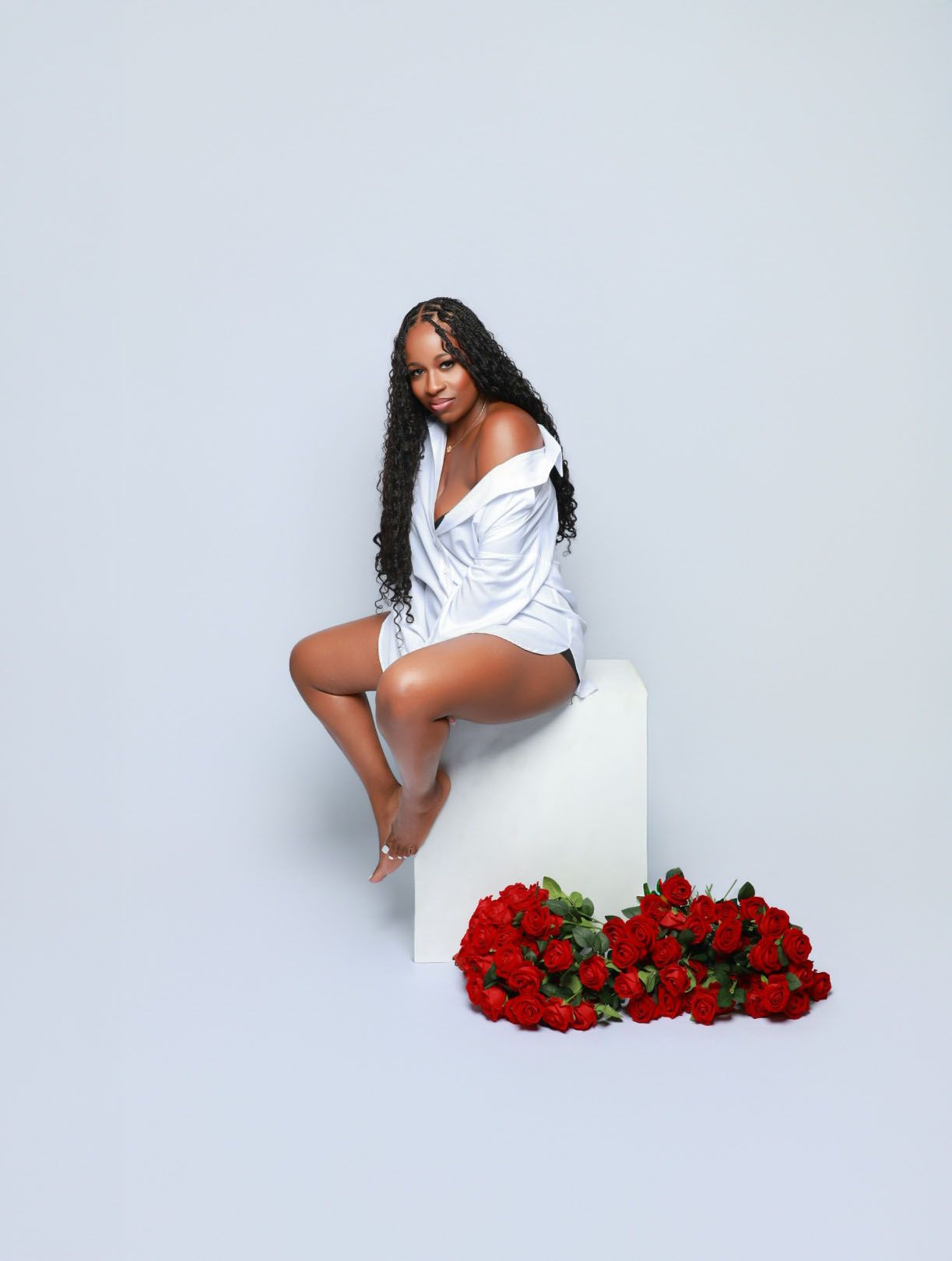 Woman in white oversized shirt sits on white cube beside red roses bouquet.