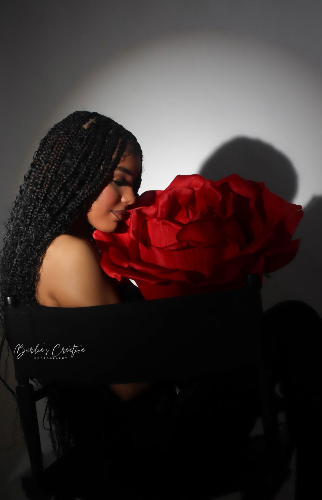 Woman with long braids smelling a large red rose in moody black and white studio photo.