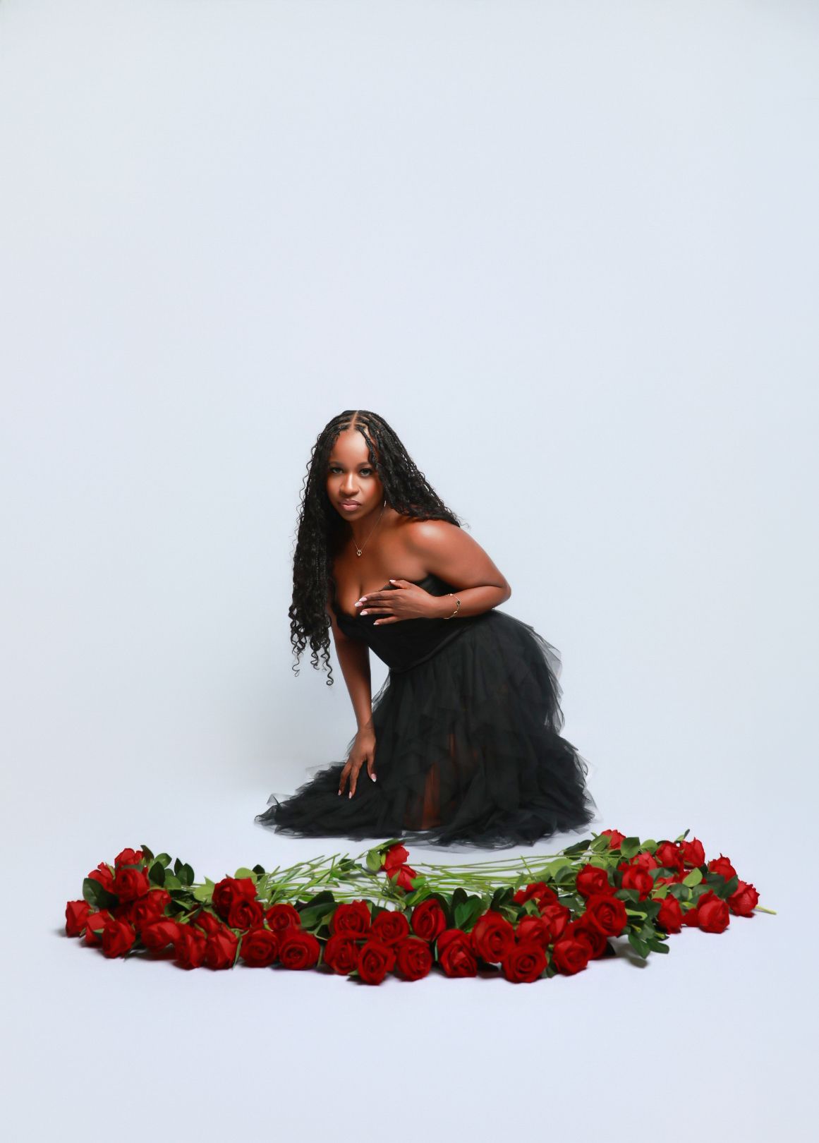 Woman in black tulle skirt kneels surrounded by red roses heart arrangement.