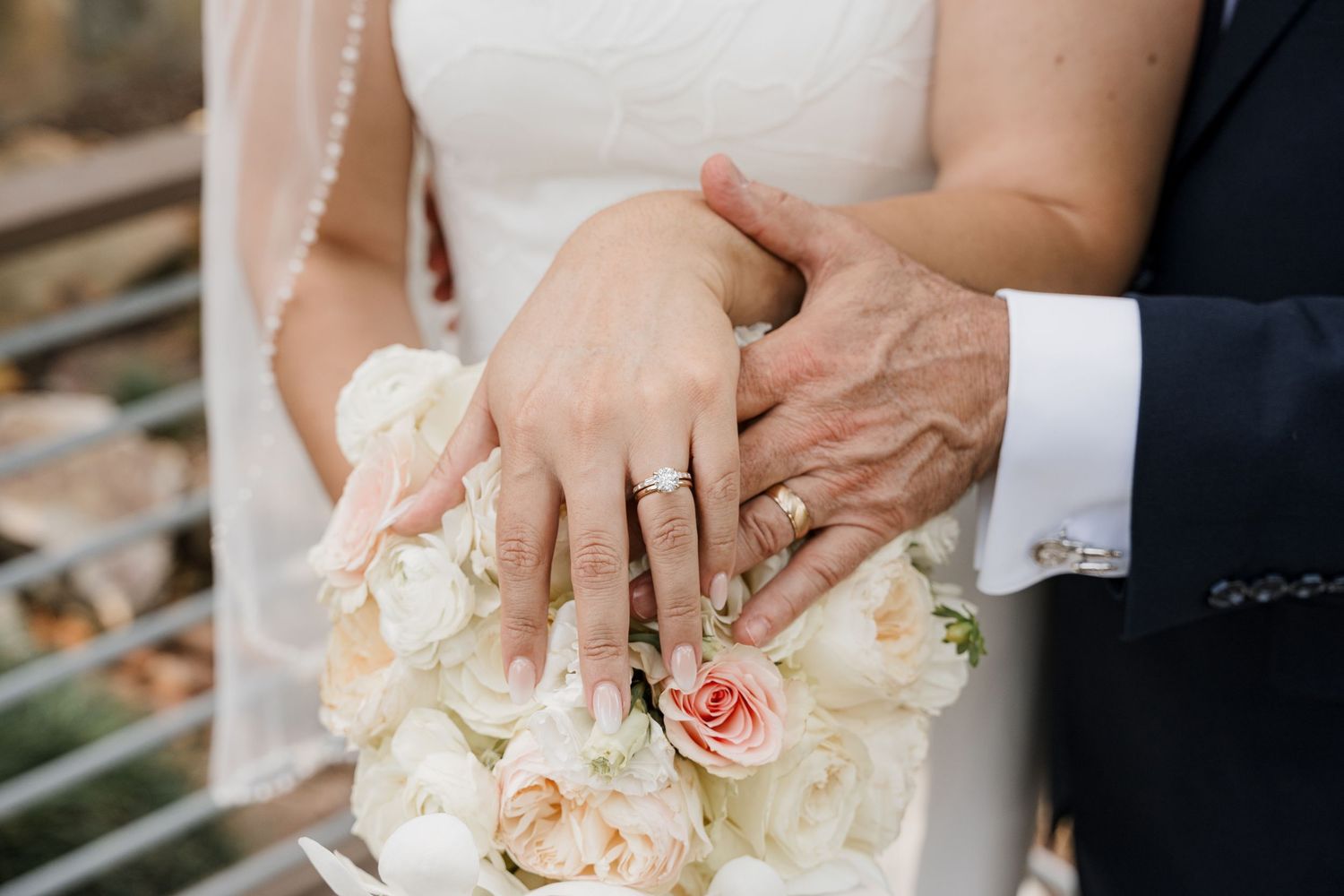 Bride and groom displaying wedding rings on hands holding white and pink rose bouquet at wedding ceremony.