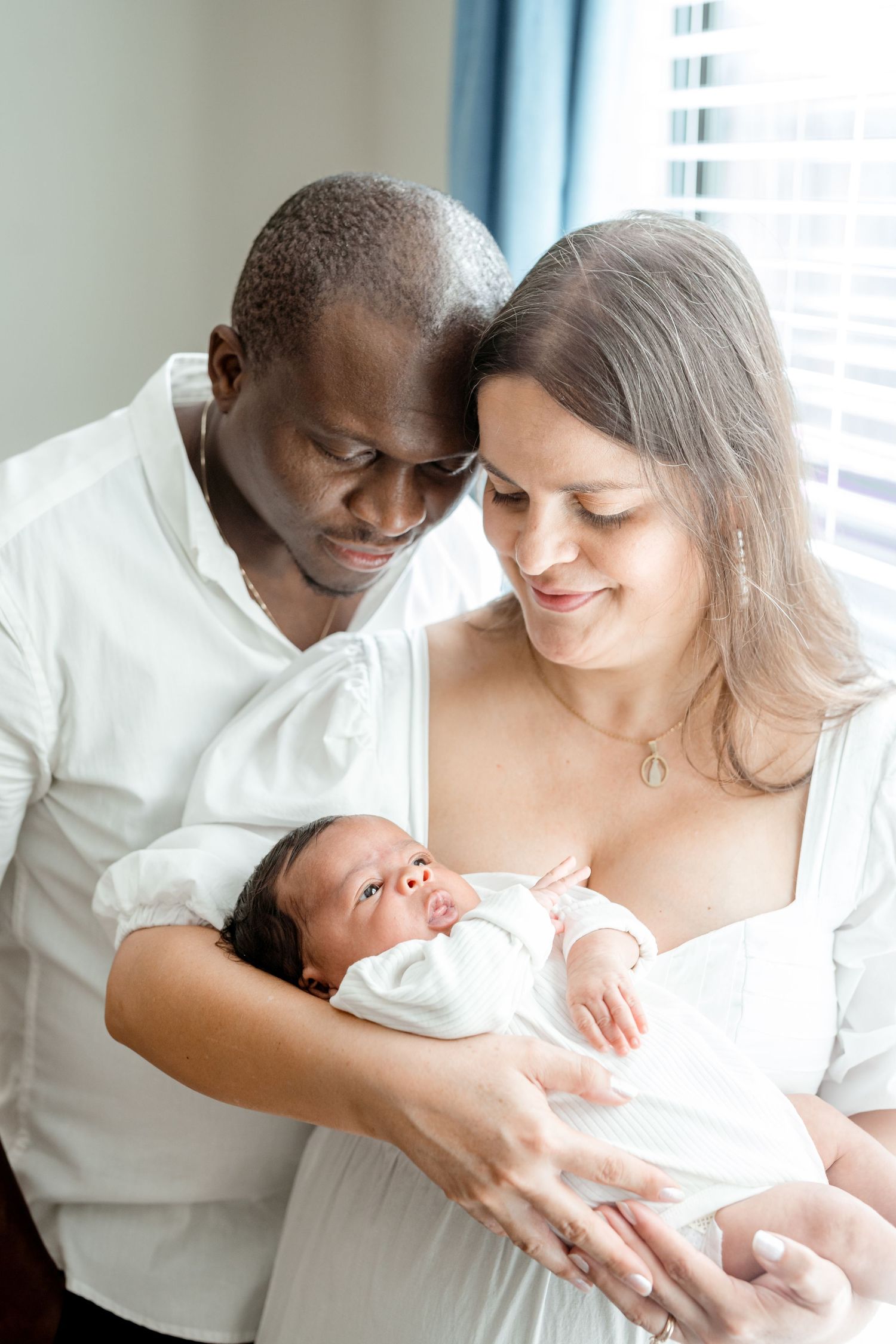 Parents in white clothing sharing tender moment with newborn baby near window.