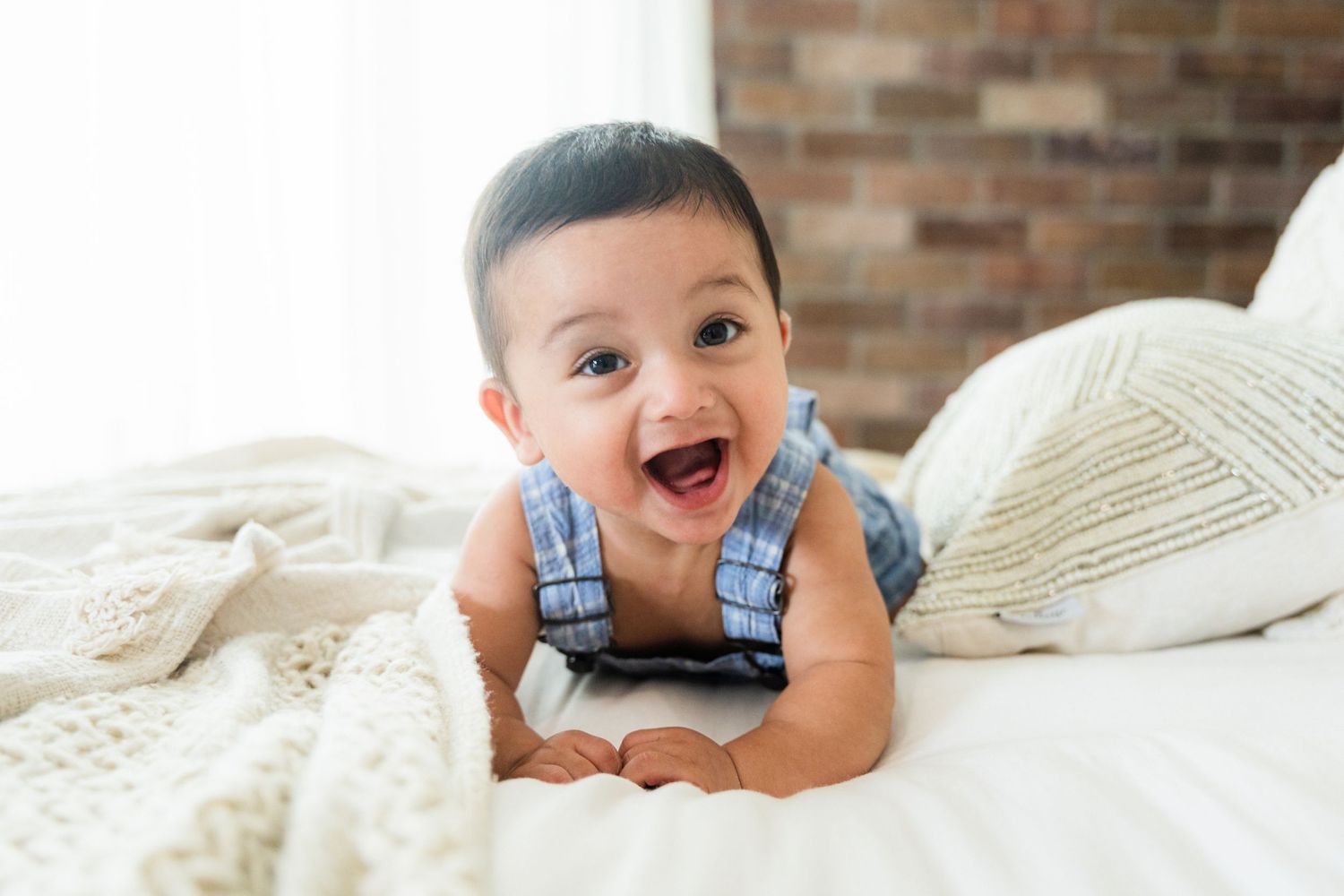 Happy baby in blue overalls lying on white knit blanket against brick wall.