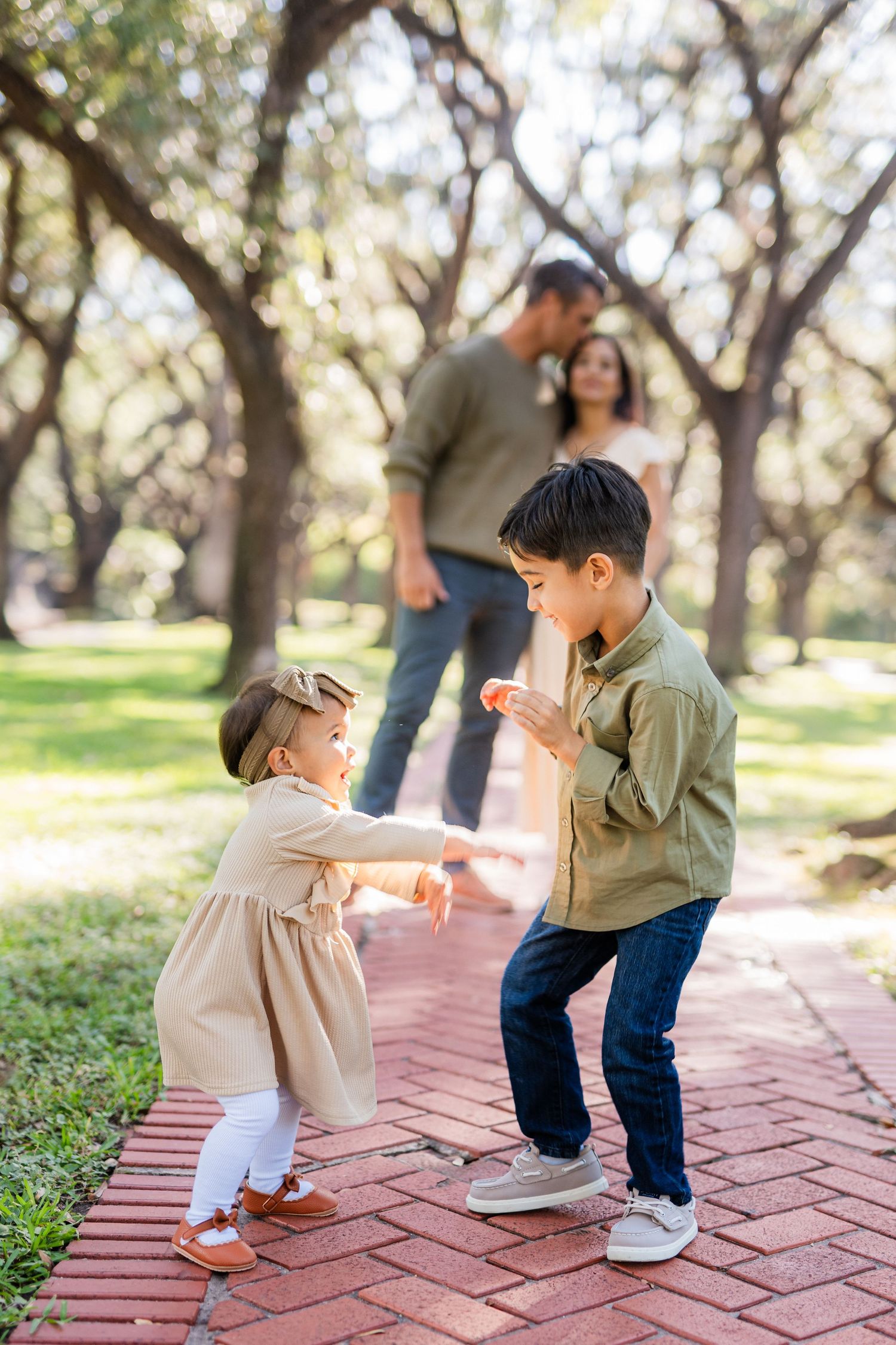 Two children playing and dancing together on a brick pathway while adults watch in the background.