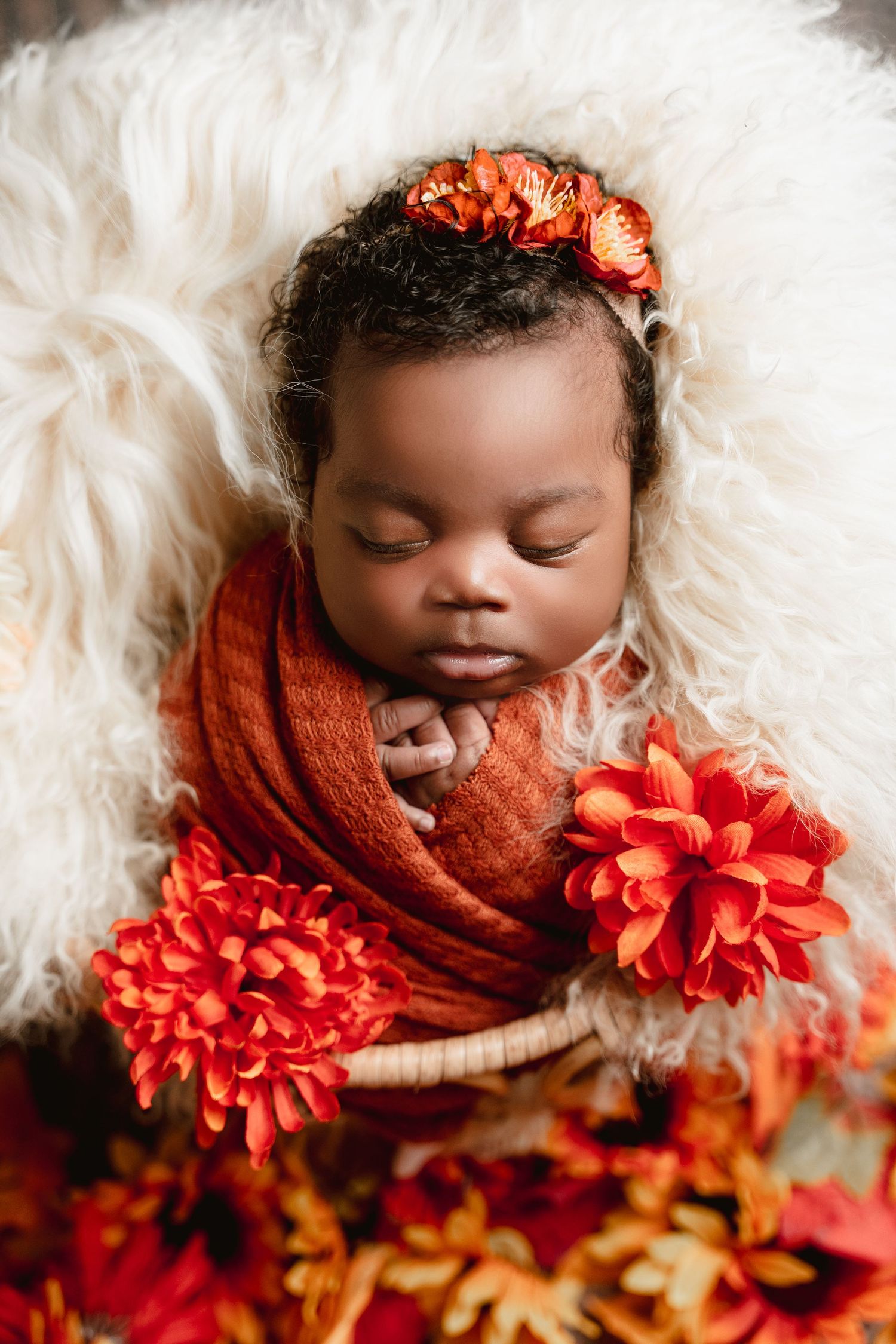 A sleeping newborn wrapped in orange fabric lies on white fur with red autumn flowers and fall leaves.
