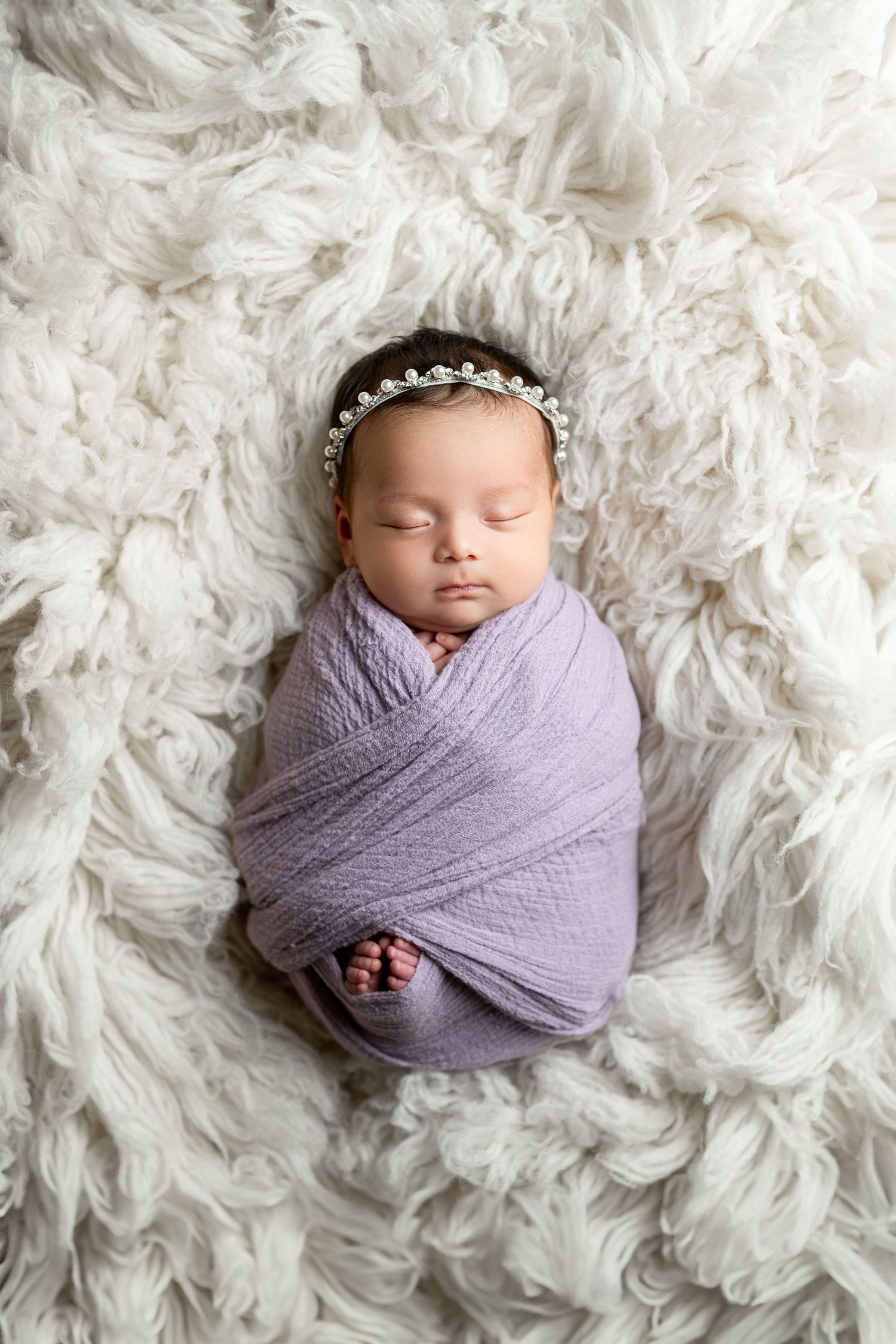 Sleeping newborn wrapped in purple swaddle on white fluffy blanket wearing crystal headband.