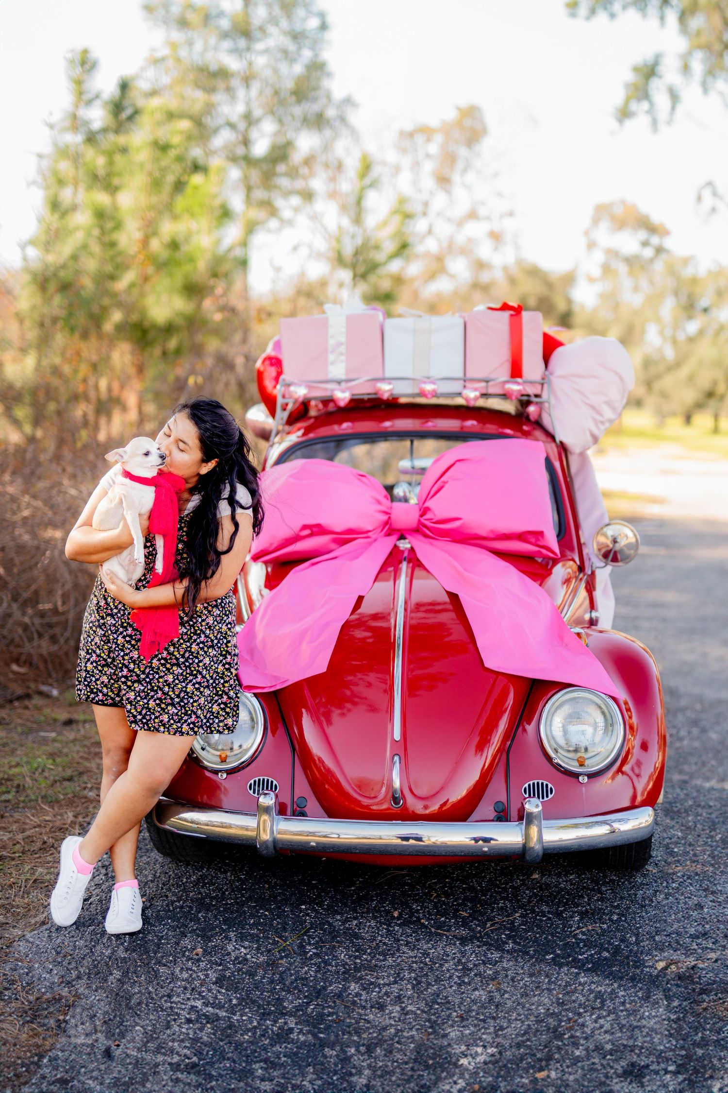 Red Volkswagen Beetle decorated with pink bow and holiday gifts on top parked on outdoor road.