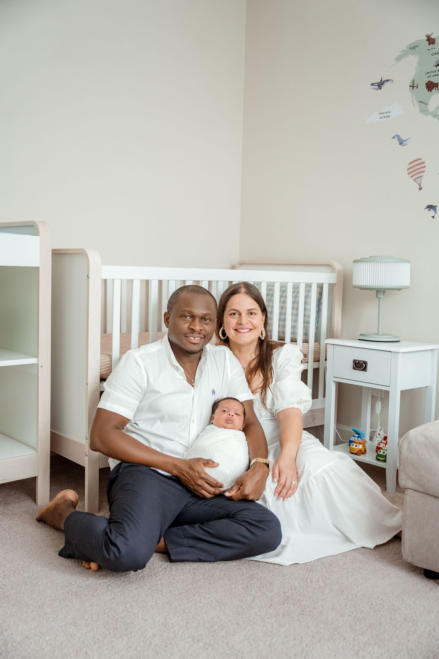 Family of three pose together in nursery with white crib and decorative wall map.