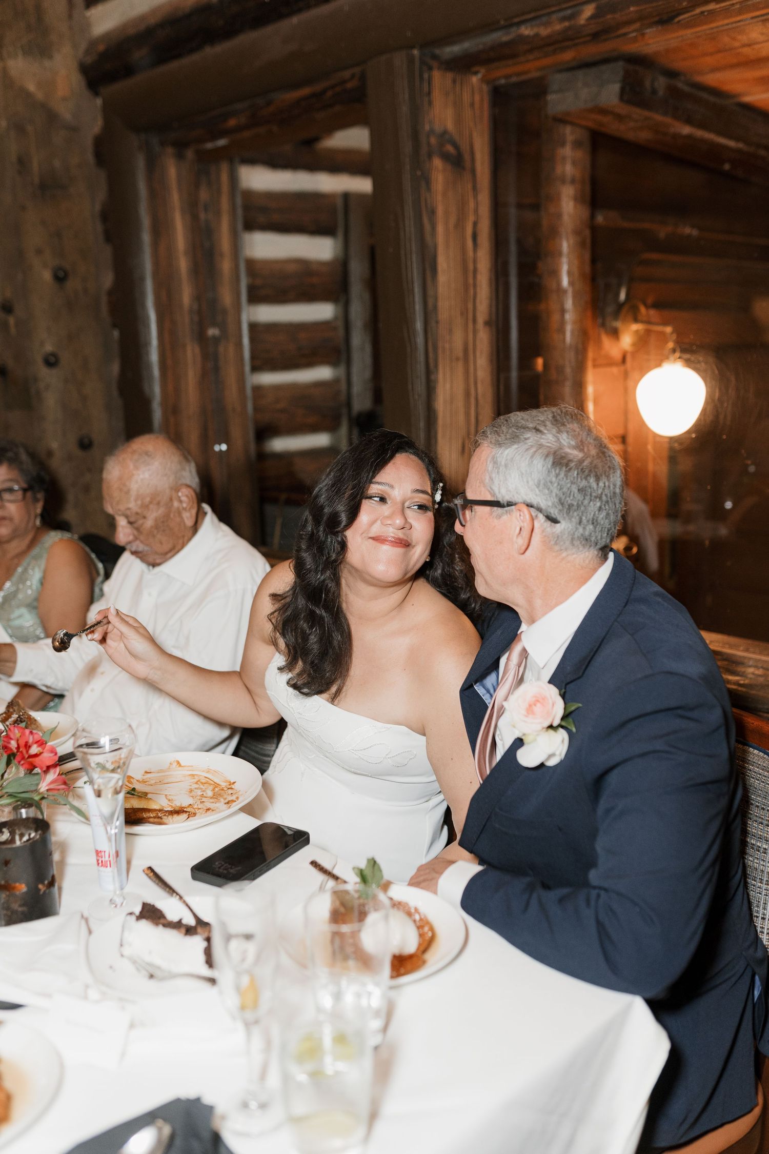 Bride in strapless gown smiles with groom in navy suit at rustic log cabin wedding reception dinner table.