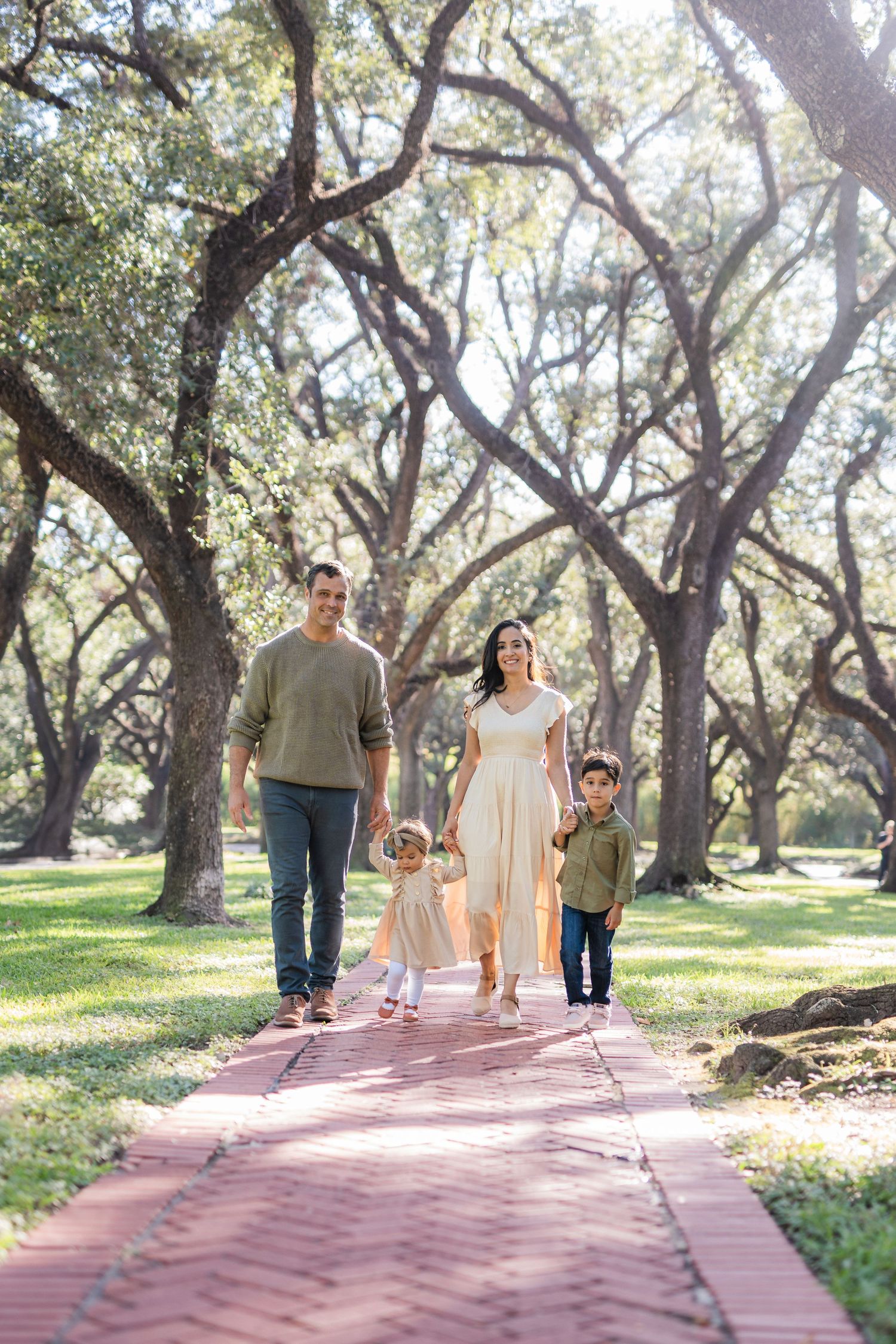 A family walking together on a red brick path through a grove of oak trees.