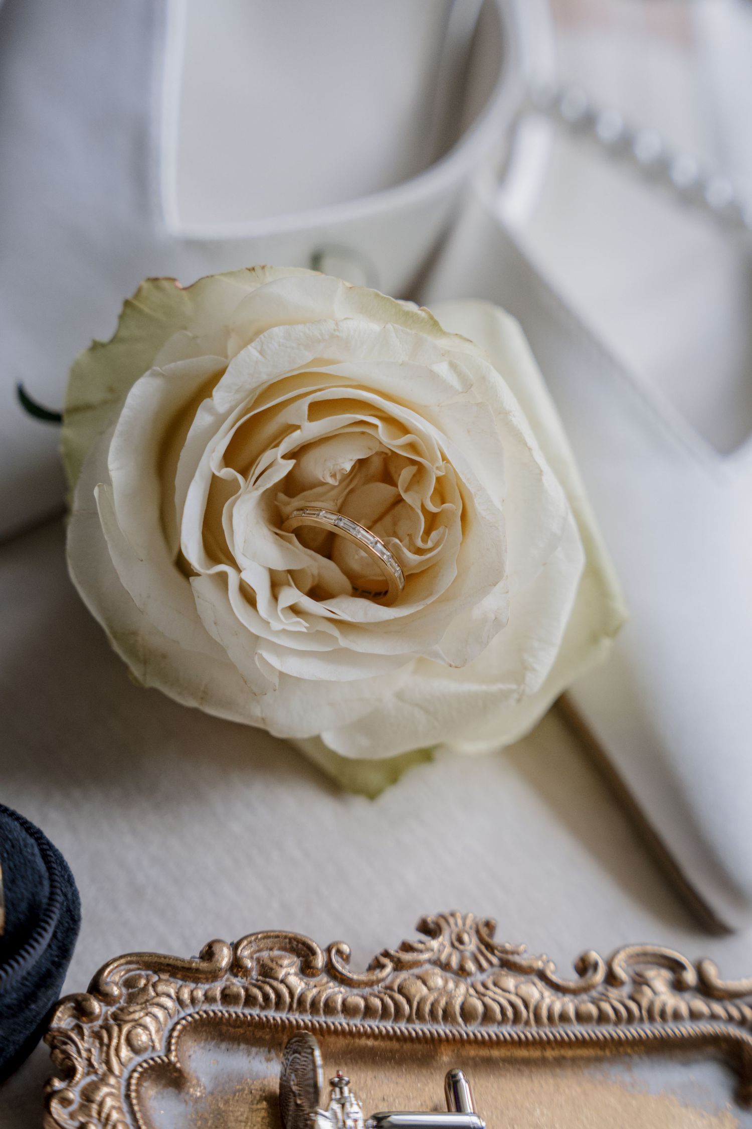 Wedding rings nestled in a white rose beside bridal heels and ornate gold frame, elegant wedding detail flat lay.