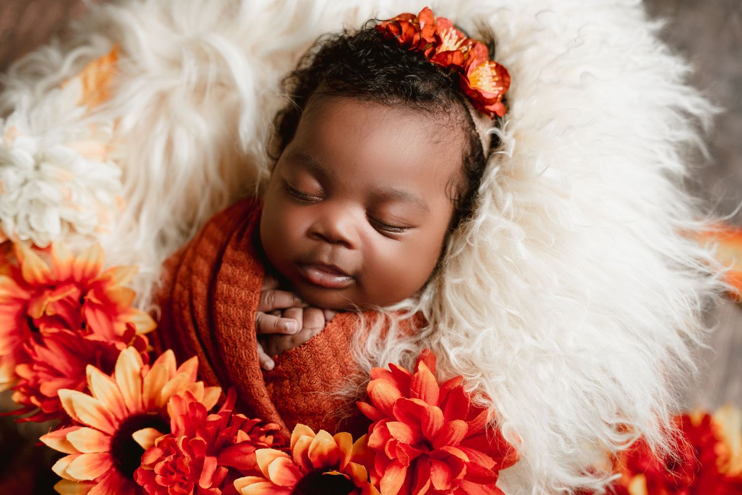 Peacefully sleeping baby wrapped in orange knit blanket surrounded by bright coral flowers on white fur background.