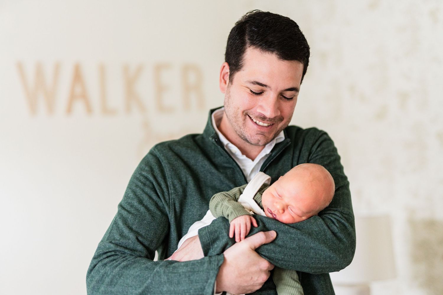 Parent wearing green sweater holds sleeping newborn with joyful expression.