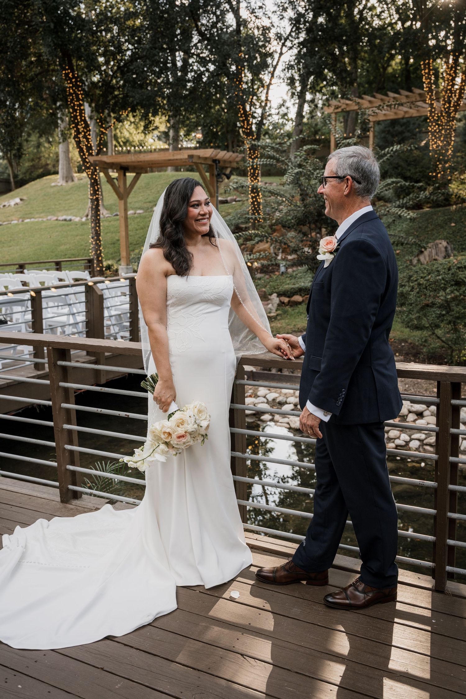 Bride in white gown holding bouquet smiles at groom in navy suit on wooden bridge at outdoor wedding venue.