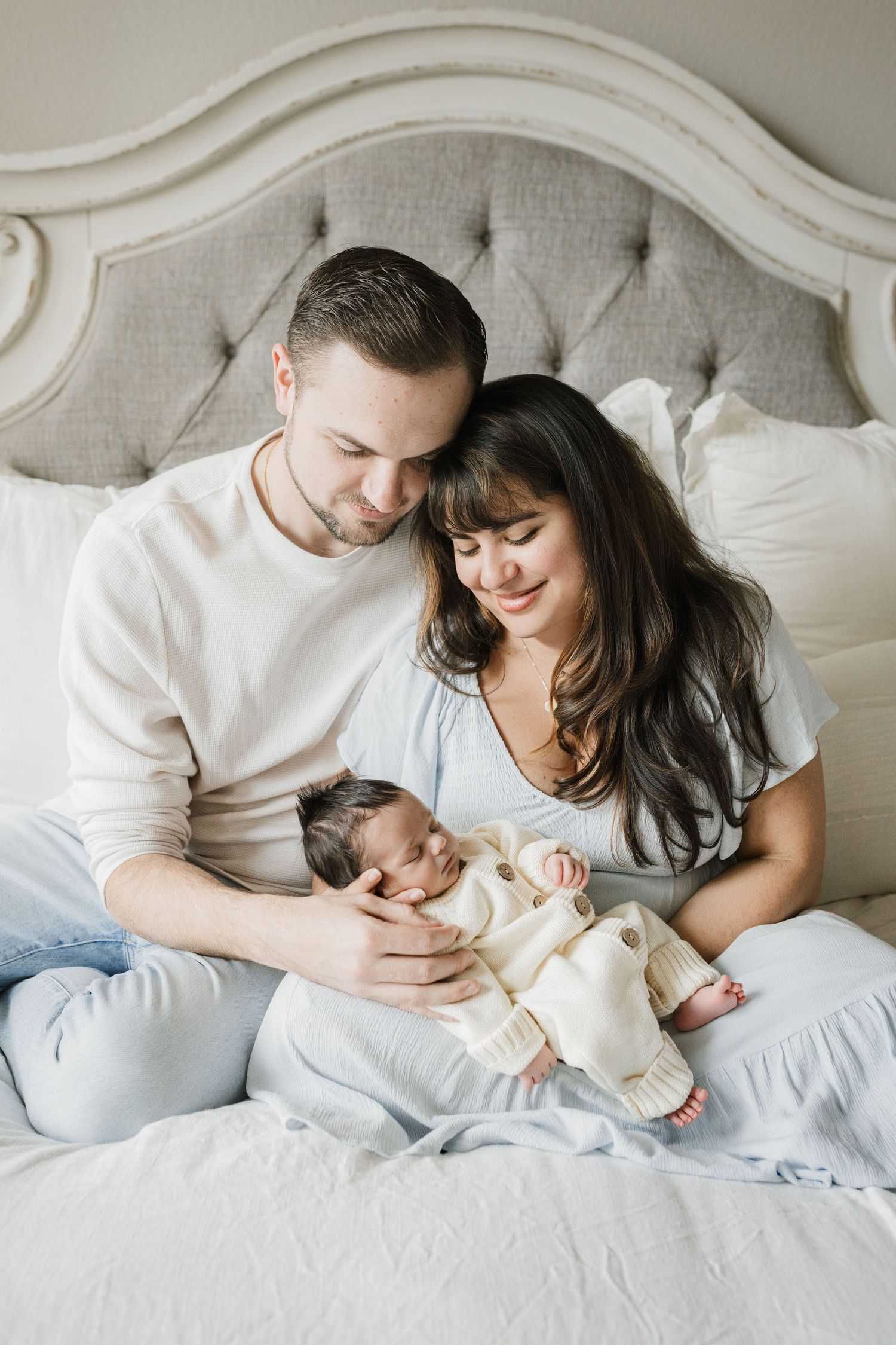 Family lying together on white bedding in cozy bedroom setting with natural light streaming in.