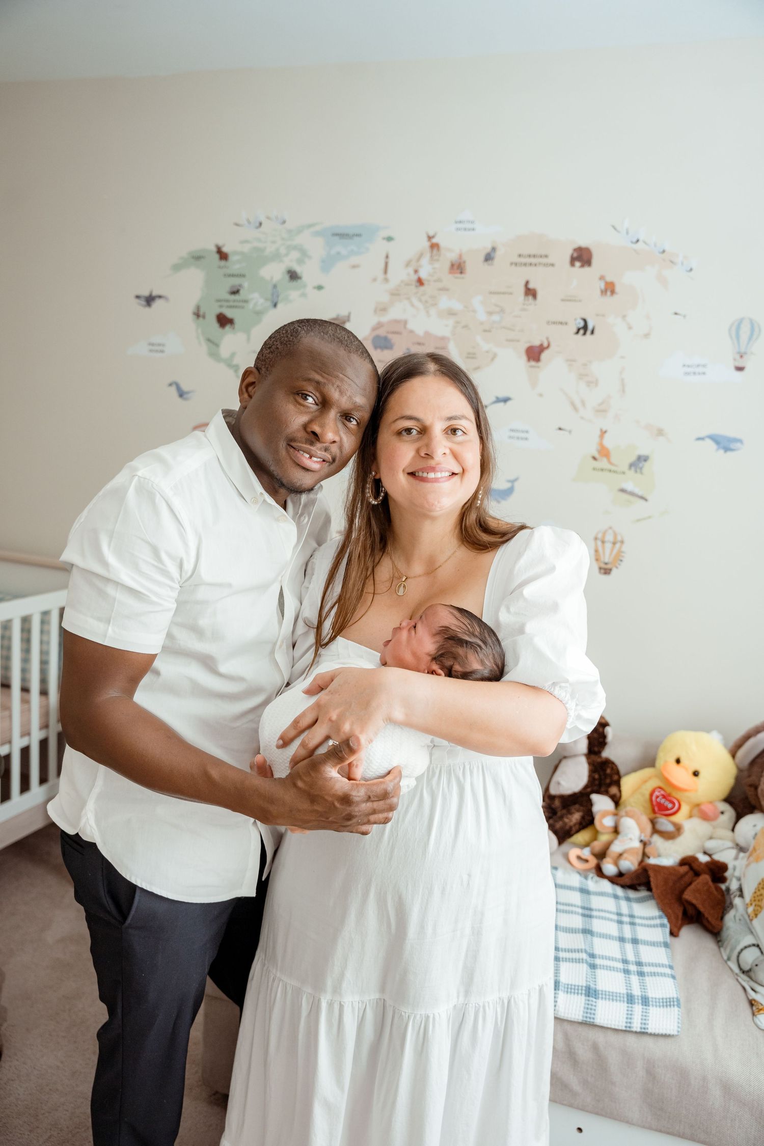 Parents in white outfits pose with newborn in nursery featuring world map wall art.