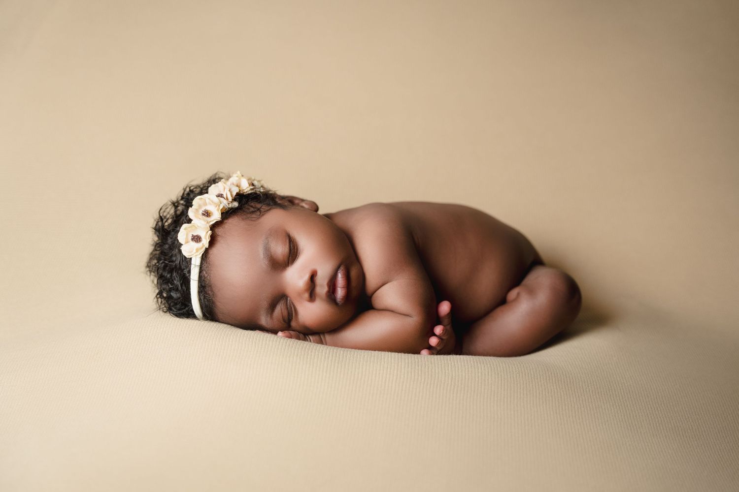 A sleeping newborn wearing a delicate flower crown lies peacefully curled up on a beige backdrop.