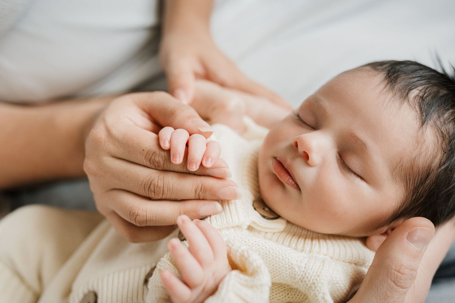 A series of images showing gentle hands holding and comforting a sleeping newborn baby wrapped in a cream blanket.