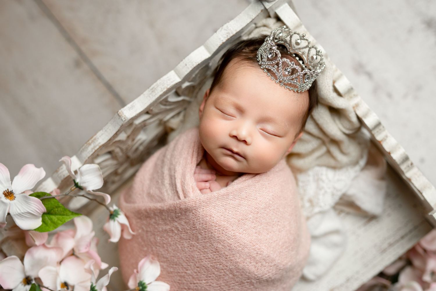 Newborn wrapped in pink blanket with crown headpiece and cherry blossoms in rustic wooden setting.