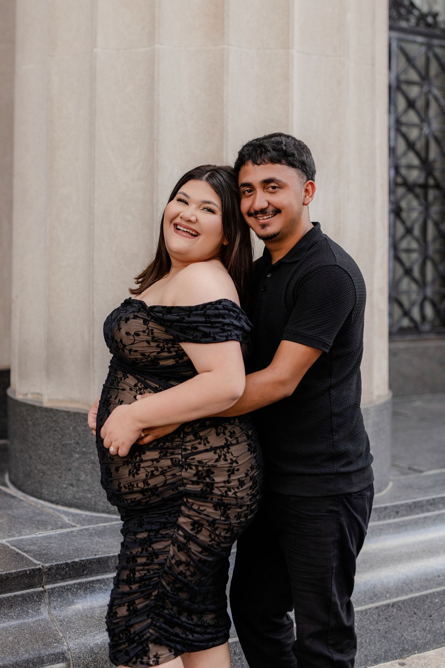 A couple embraces in front of stone columns, with the expectant mother wearing a black lace off-shoulder dress.