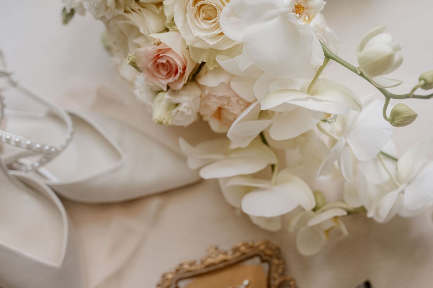 Elegant bridal flat lay with white orchids, blush roses, pearl heels, and a vintage gold mirror on soft white fabric.