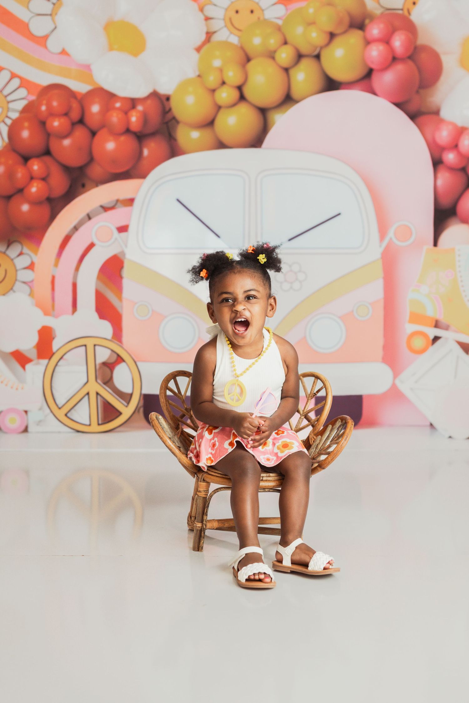 Joyful child sits in wooden chair against colorful balloon backdrop wearing tutu dress and white sandals.