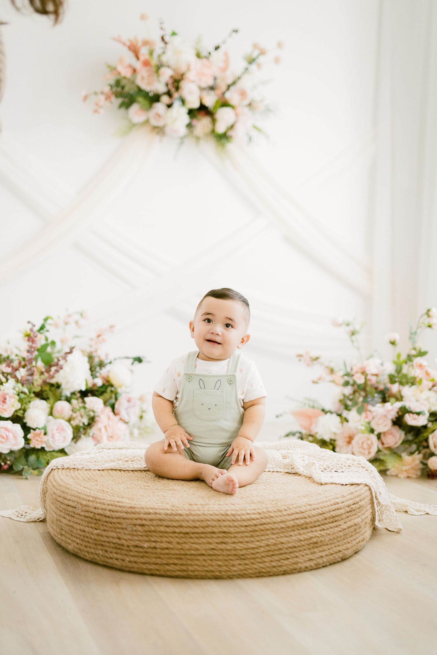 Baby in mint green overalls sits on round jute pouf with floral arrangements in soft pink and white tones.