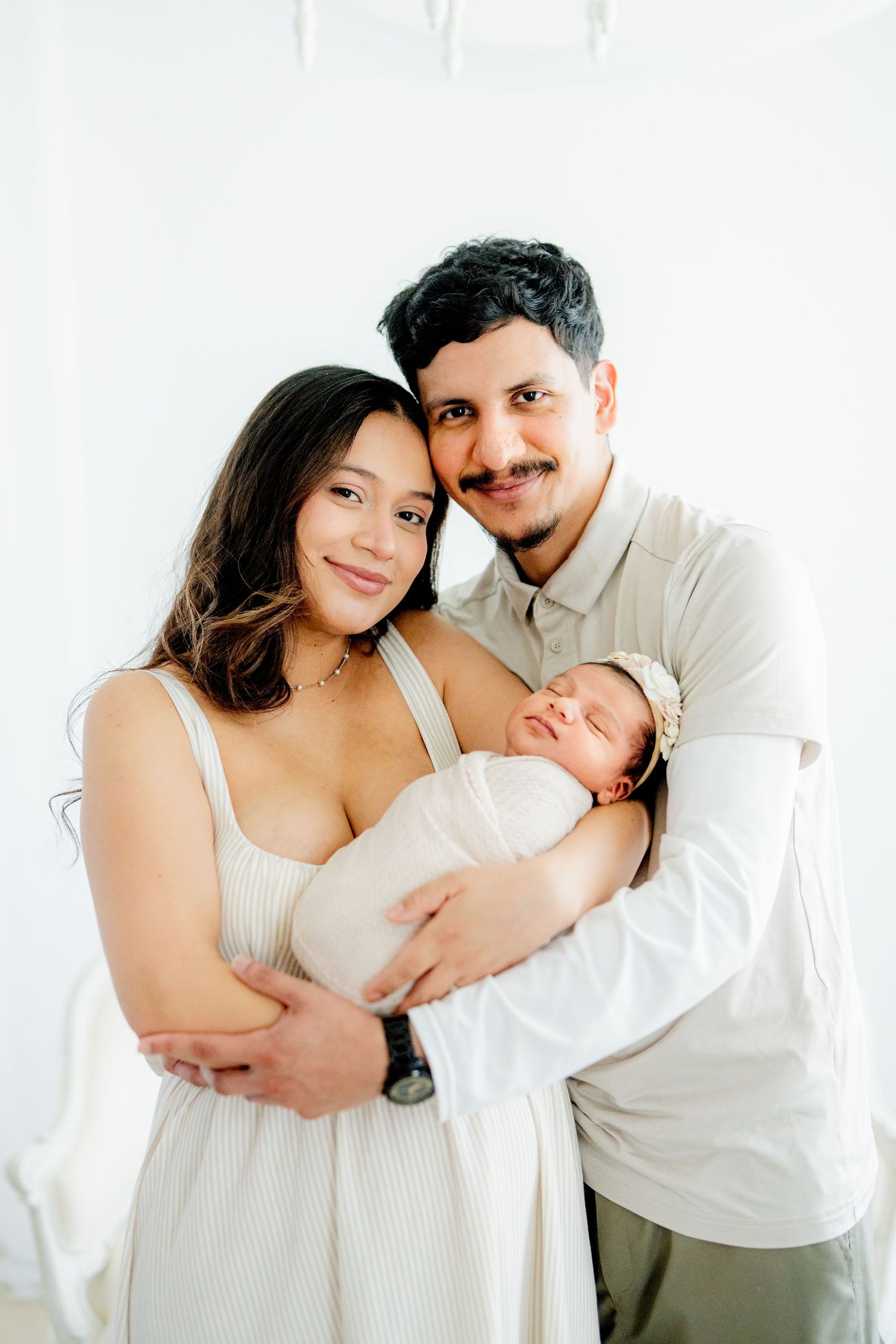 A sleeping newborn baby swaddled in white fabric being cradled by adults in a bright studio setting.