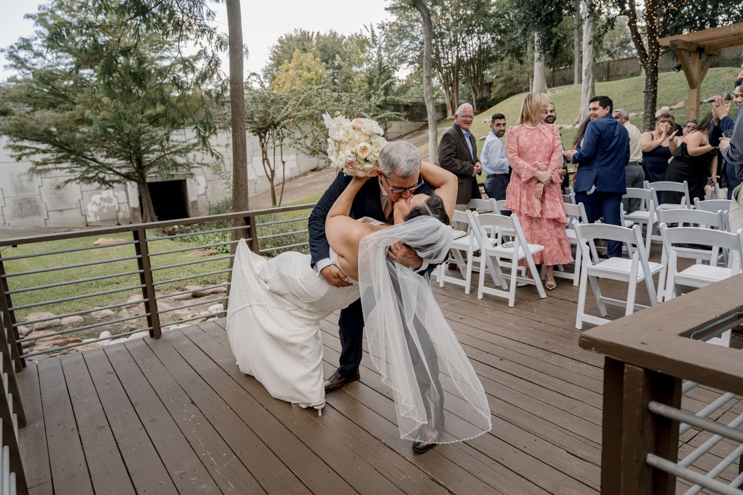 Groom dips bride for a kiss on wooden deck during outdoor wedding ceremony with guests seated nearby.