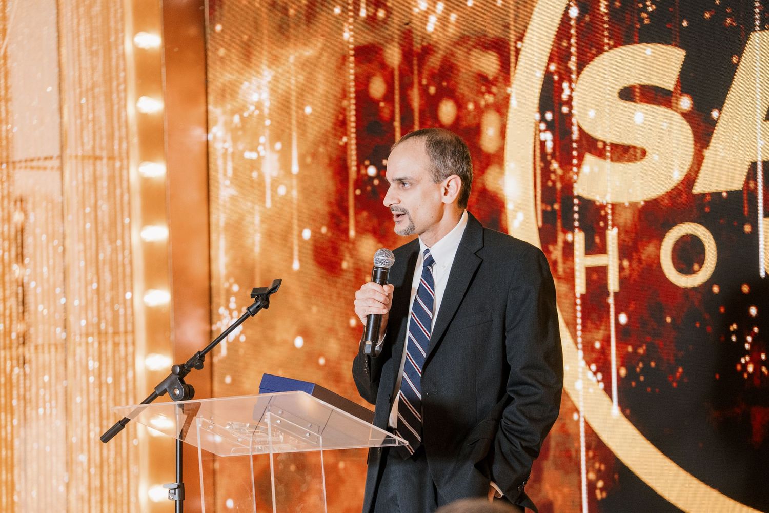 Man in suit speaking at podium with microphone at formal awards ceremony with golden bokeh backdrop.