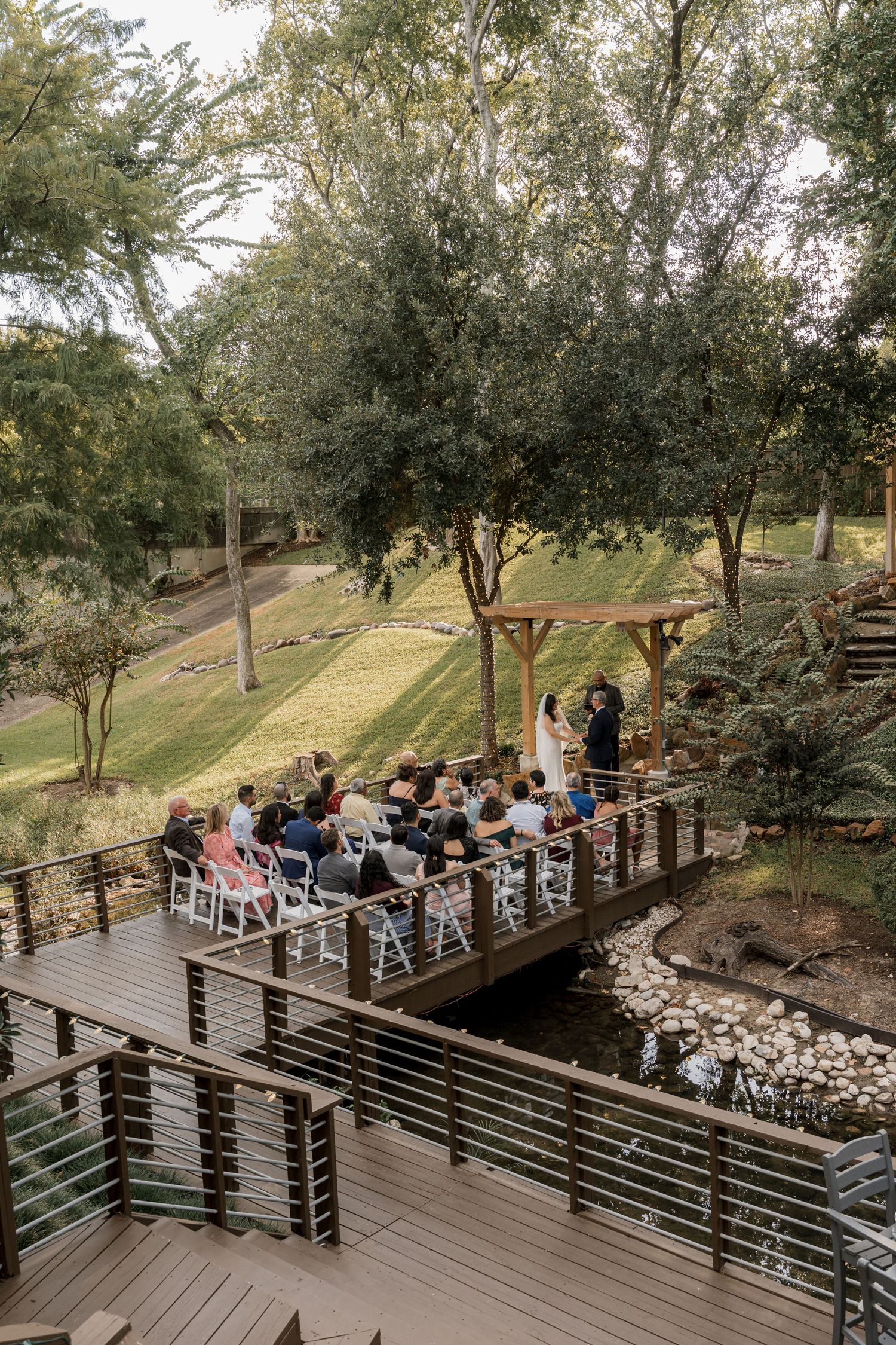 Outdoor wedding ceremony on a wooden bridge deck surrounded by lush trees and a rocky stream at a scenic garden venue.