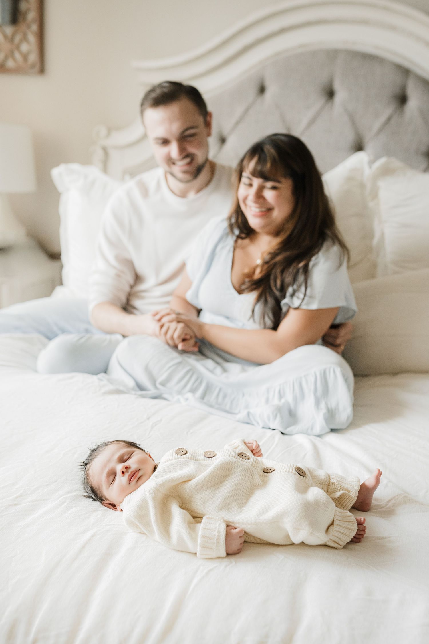 A couple sits together on a white bed with their newborn baby in a cream-colored outfit lying in front of them.